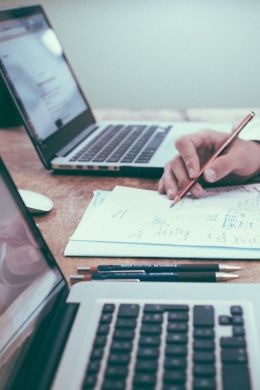 Zwei Laptops und Unterlagen, in denen eine Person schreibt